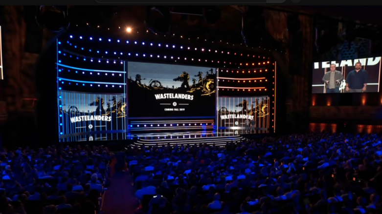 Two guys stand in front of a Fallout 76 Wastelanders screen and a crowd of people